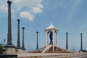 Home 3 A photograph of the Mahatma Gandhi statue under a white canopy, surrounded by eight tall, carved granite pillars on the Seaside Promenade in Pondicherry, under a blue sky with clouds.