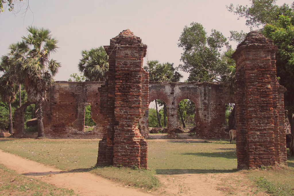 The brick ruins of the 18th-century French Jesuit Mission House at the archaeological site of Arikamedu near Pondicherry, surrounded by dry grass and palm trees under a clear sky.