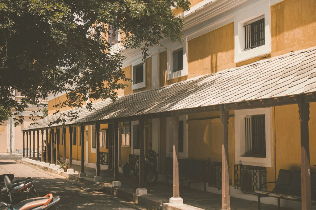 long, mustard-yellow colonial building in White Town, Puducherry, featuring a white-trimmed roof, wooden pillars supporting a tiled awning, and barred windows. A large tree with lush leaves overhangs the left side of the building, casting shadows. Parked motorbikes are visible in the foreground.