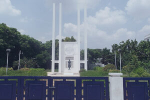 The French War Memorial in Pondicherry, a white monument with tall columns and a bronze statue of a soldier, located on Goubert Avenue (Promenade) and surrounded by a blue fence and greenery under a cloudy sky.