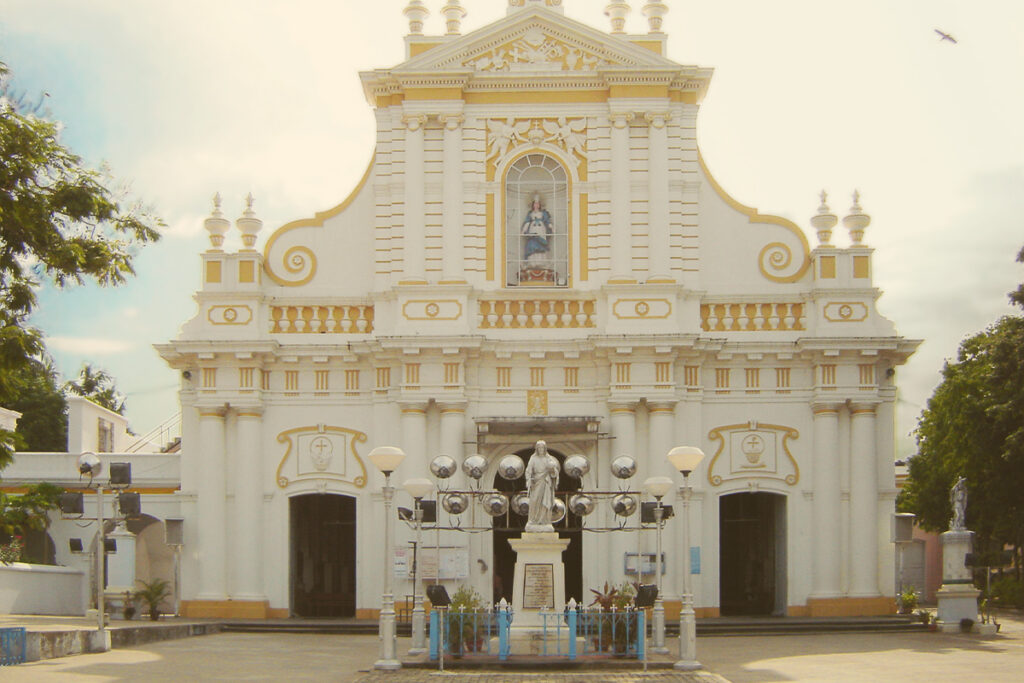 The facade of the Immaculate Conception Cathedral (Samba Kovil) in Puducherry, showcasing its Portuguese-style architecture, white exterior with yellow trim, a statue of Mother Mary in a central niche, and a statue of Jesus Christ in front of the entrance under a cloudy sky.