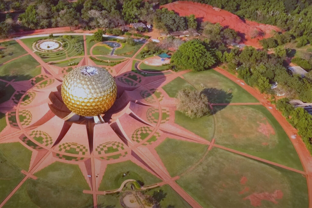 A stunning aerial photograph of the Matrimandir, a giant golden globe structure, situated at the heart of Auroville and surrounded by the intricately designed Peace Gardens and lush greenery.
