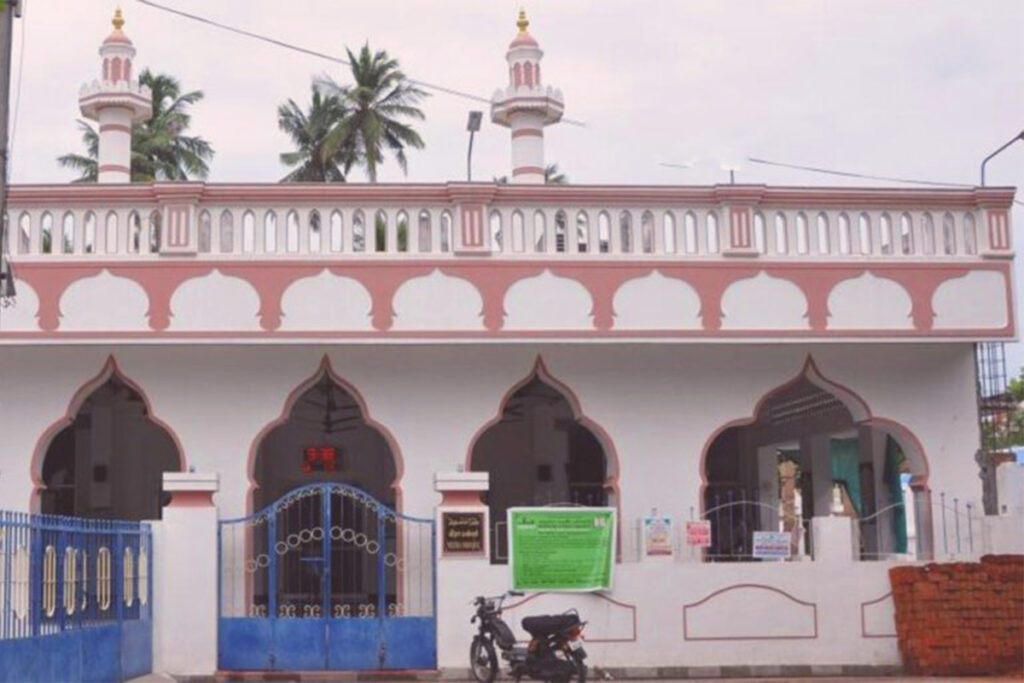 The exterior facade of the Meeran Mosque (Meerapalli) in Pondicherry, featuring a white structure with distinct pink-trimmed Islamic arches, a blue entry gate, and small minarets against a backdrop of palm trees.