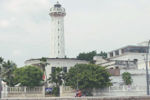 A view of the historic Old Lighthouse in Pondicherry, a tall white tower standing behind a sandy beach and surrounded by trees under an overcast sky.