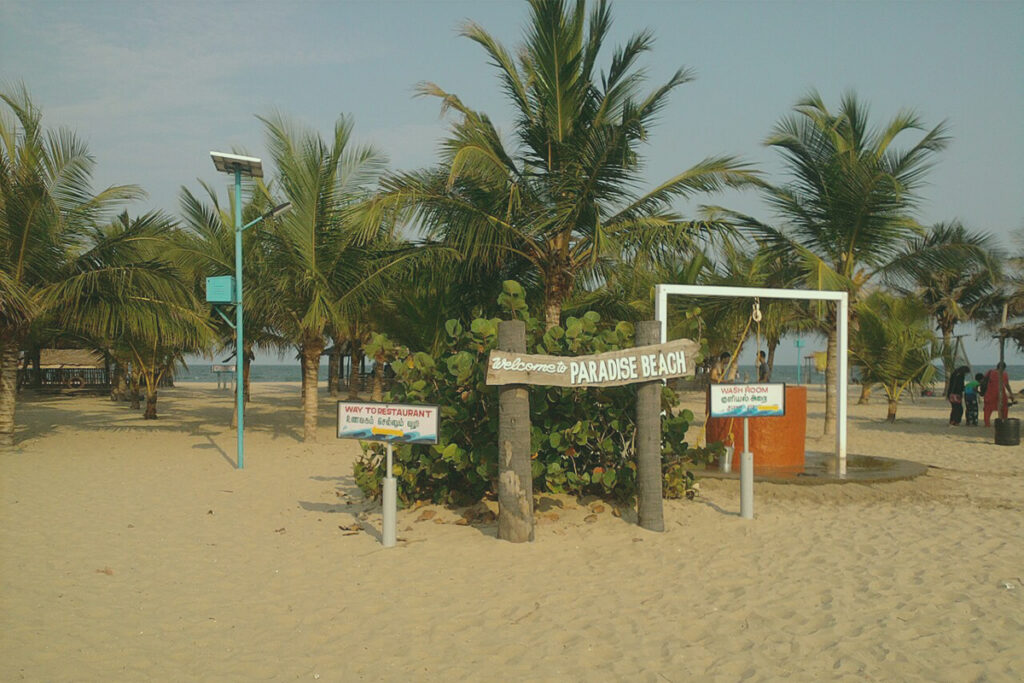 A view of the sandy entrance to Paradise Beach in Pondicherry, featuring a wooden sign that reads "Welcome to PARADISE BEACH", another sign for the restaurant, and several palm trees under a sunny sky.