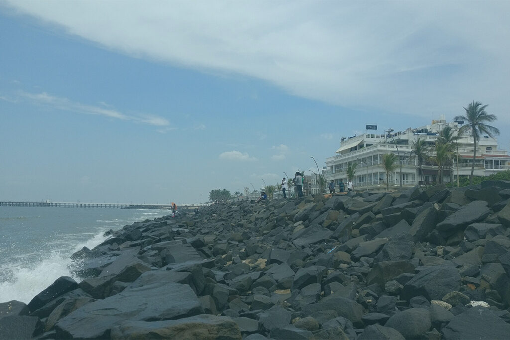A wide-angle view of the Seaside Promenade in Pondicherry, showing the rocky shoreline, the Bay of Bengal with crashing waves, and a row of colonial-style buildings under a blue sky with clouds. A long pier stretches into the sea in the distance.