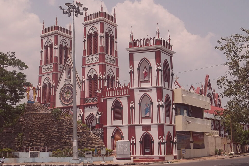 The exterior of the Basilica of the Sacred Heart of Jesus in Puducherry, showcasing its distinct red and white Gothic Revival architecture, twin spires, a large rose window, and a rock grotto with a statue of Jesus to the left under a partly cloudy sky.