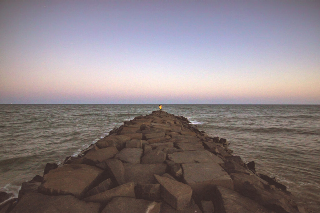 A breathtaking sunrise over Serenity Beach in Pondicherry, where a long rock pier extends into the Bay of Bengal. A lone person stands at the far end of the pier, watching the horizon as the sky transitions from purple and blue to soft pink and orange.
