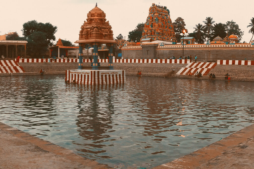 A panoramic view of the Sri Gokilambal Thirukameswarar Temple (Villianur Temple) in Puducherry, featuring its large sacred temple tank with steps leading down to the water, and multiple towering, intricately carved gopurams under a partly cloudy sky.