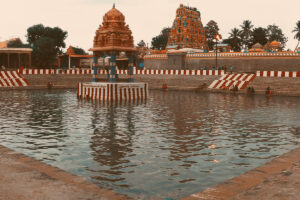 A panoramic view of the Sri Gokilambal Thirukameswarar Temple (Villianur Temple) in Puducherry, featuring its large sacred temple tank with steps leading down to the water, and multiple towering, intricately carved gopurams under a partly cloudy sky.