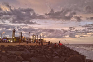 People enjoying a dramatic sunset over the Bay of Bengal from the rocky shore of Rock Beach on the Seaside Promenade in Pondicherry, India, with the Gandhi Statue and lighthouse in the background.
