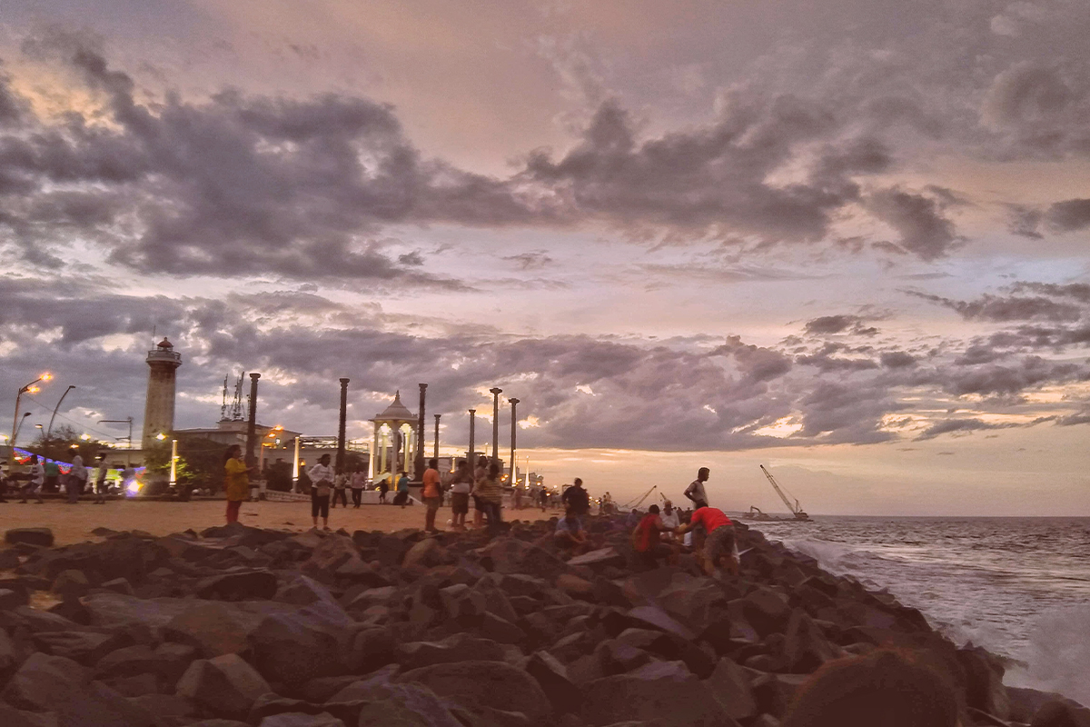 People enjoying a dramatic sunset over the Bay of Bengal from the rocky shore of Rock Beach on the Seaside Promenade in Pondicherry, India, with the Gandhi Statue and lighthouse in the background.